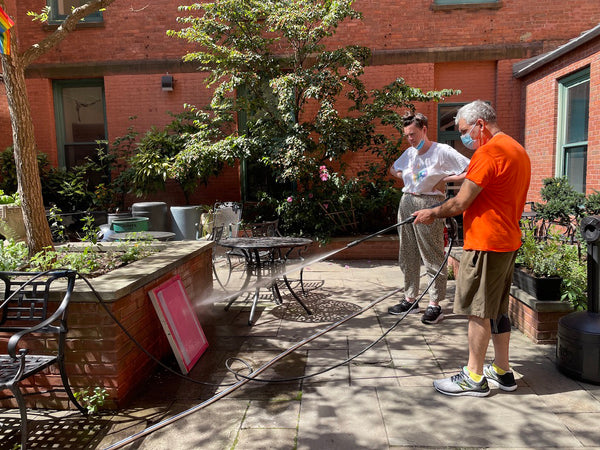 Roger Jones is pressure washing a silk screen while Natalie Gaimari watches him. She has her arms on her hips. They are in a sunny red brick courtyard with  a tree in the background.